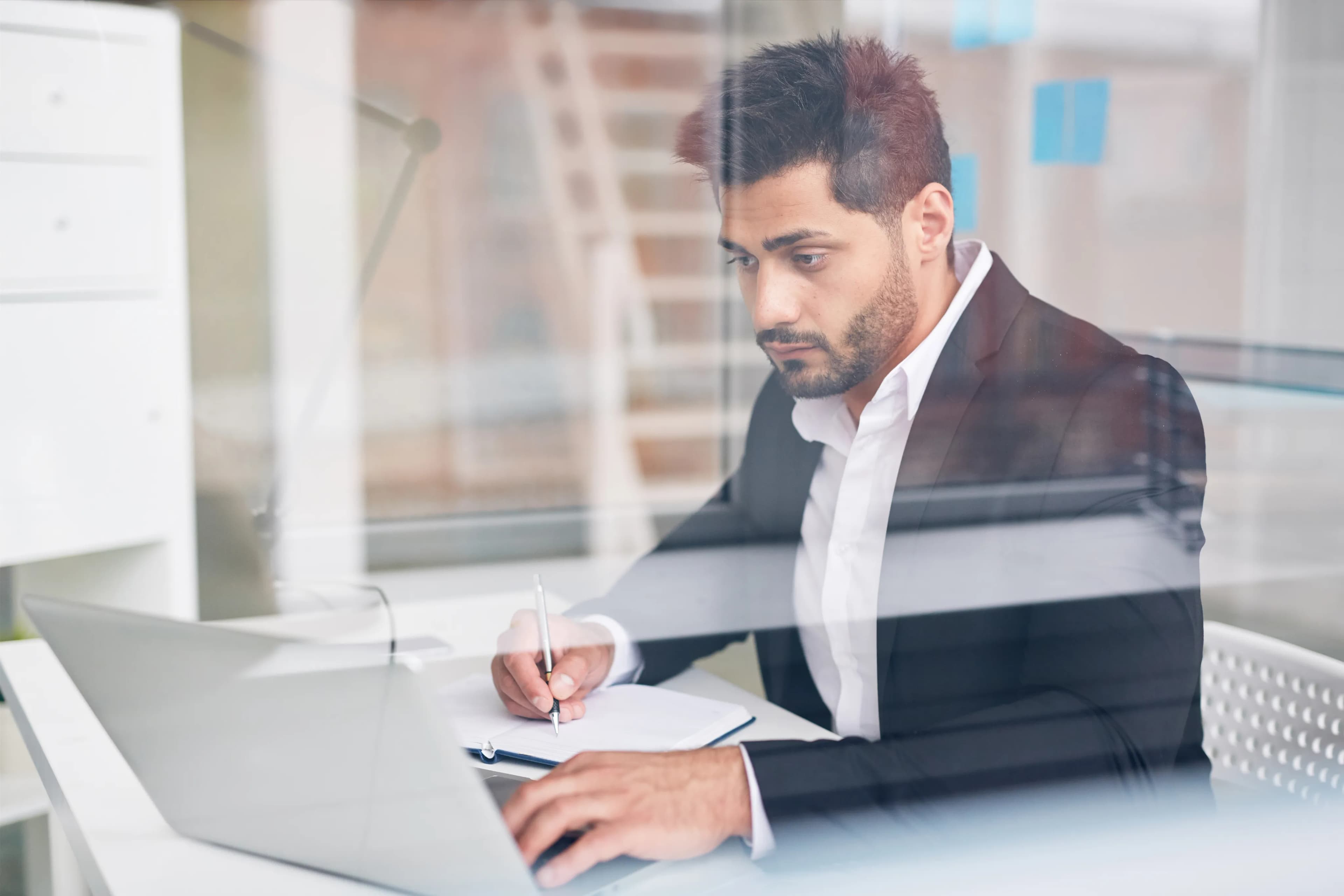 Professional man in a suit works at a desk with a laptop and notebook, symbolizing ethical AI innovation and responsible decision-making in a modern, transparent office environment. The scene highlights focus, accountability, and fairness in technology-driven workplaces.